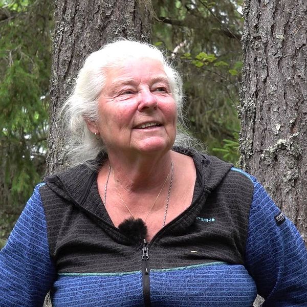 Kvinna med uppsatt grått hår och blåsvart tröja. Hon sitter i skogen och blickar åt höger.