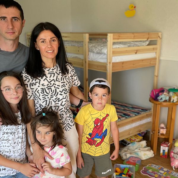 Två föräldrar står med sina tre barn i ett sovrum med leksaker på golvet.