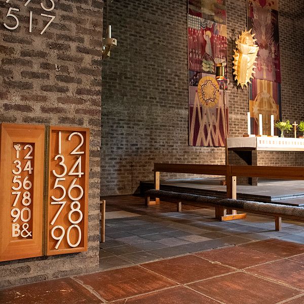 Interiör i en kyrka med altartavlor mot en tegelvägg, en stol, kor och altare med duk och stearinljus.