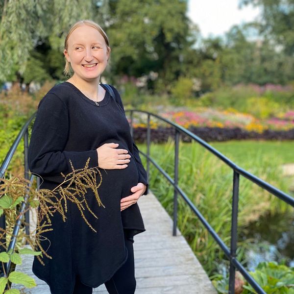 Gravid leende kvinna i svart står på bro i grön park