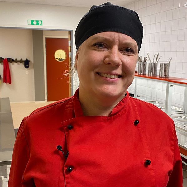 En kökschef med röd kockjacka och kolsvart kockmössa som jobbar i en skolmatsal i Gustafs.