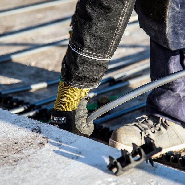 byggarbetsplats, byggnadsarbetare som installerar golvvärme i ett betonggolv, grova kängor, jobbarkläder och handskar. man ser bara fötter, ben och en hand, arbetar framåtböjd nere på golvet.