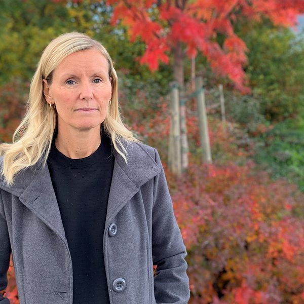 Linda Larsson, enhetschef på individ- och familjeomsorgen i Skellefteå kommun ,står utomhus. i bakgrunden syns träd i olika höstfärger. Hon är blond och klädd i en svart tröja och grå kappa.