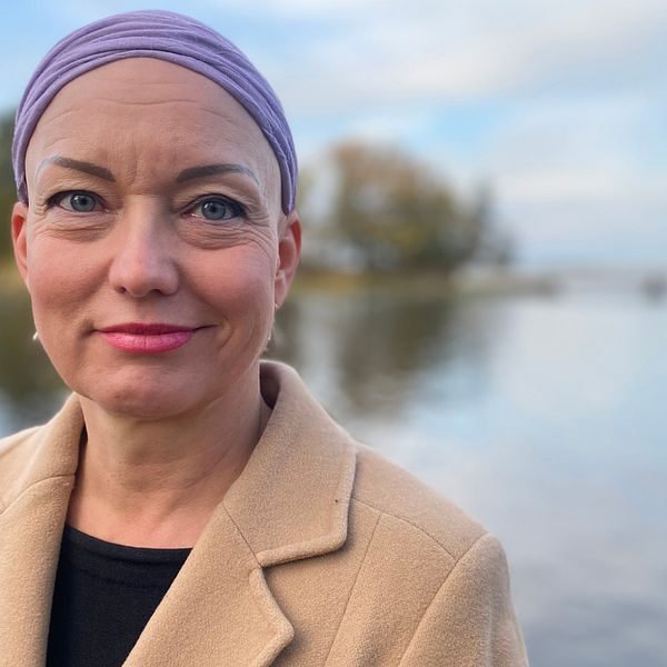 Bild på Carola Forssell, deltidsboende i Mälarbaden utanför Eskilstuna. Hon tittar in i kameran. På sig har en beige kappa med en svart tröja under. På huvudet har hon en lila sjal. Hon har rosa läppstift.