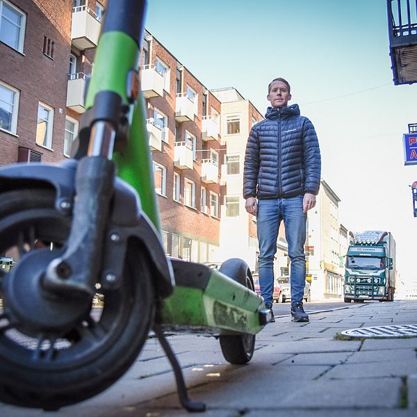Johan Larsson – en man i jeans och dunjacka – står bakom en elsparkcykel parkerad mitt på en trottoar.