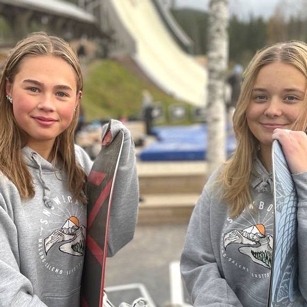 Två tonårstjejer som står och hänger på var sin snowboard utomhus, det är barmark och i bakgrunden skymtar en konstgjord skidbacke.
