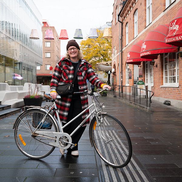 Helbild på kvinna, blont hår, brun mössa, glasögon, kappa i i rött och vitt, står och håller sin cykel på en gågata med grå stenplattor.