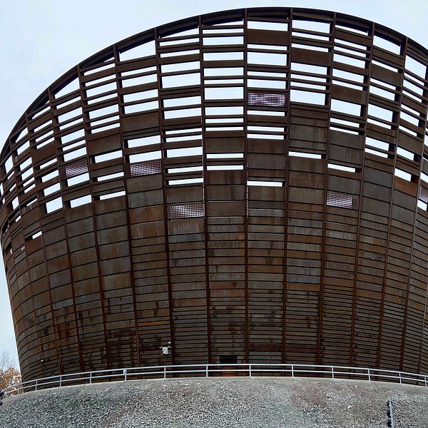 En modern, rund byggnad med ett öppet träverk som liknar en stor korg, belägen på en kulle omgiven av träd och naturlandskap. Byggnaden har en unik design med stora öppningar som släpper in ljus.