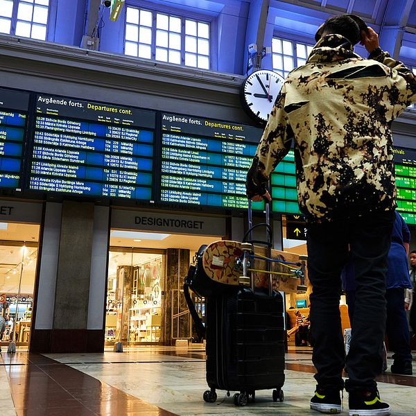 En kille står med sin resväska och tittar upp mot en digital tavla med alla tågens avgångar och ankomster.