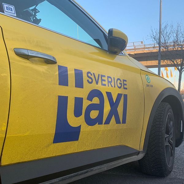 På bilden syns en taxibil från företaget Sverigetaxi. Himlen är blå och till höger i bilden i bakgrunden syns en bro.