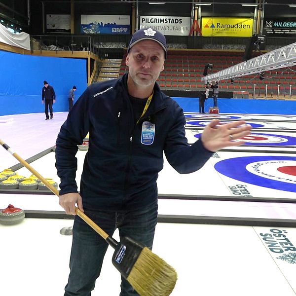Marcus Olovsson står på en curlingbana med en sop i ena handen. I bakgrunden syns en tom läktare och personer som jobbar med förberedelser inför EM i curling.
