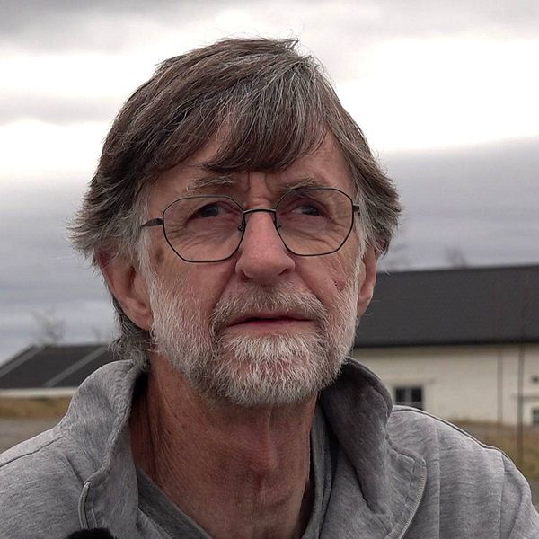 Russ Murtagh, en man i 60-årsåldern med grått hår och grått skägg och glasögon sitter på huk framför ett vitt hus med natur i bakgrunden.