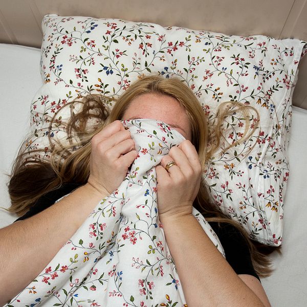 En halvbild, tagen uppifrån. En blond, ljushyad, kvinna ligger i en säng och gömmer ansiktet under ett blommigt täcke.