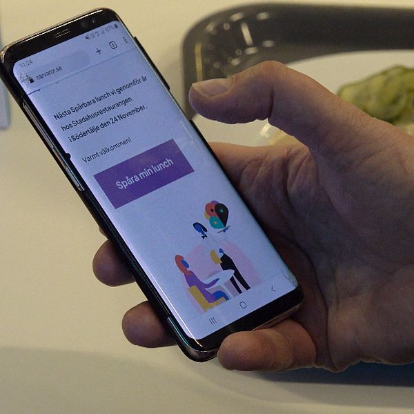 Hand som håller en mobiltelefon. På telefonen syns en ruta med texten ”spåra min lunch”. Tallrik med köttbullar i bakgrunden