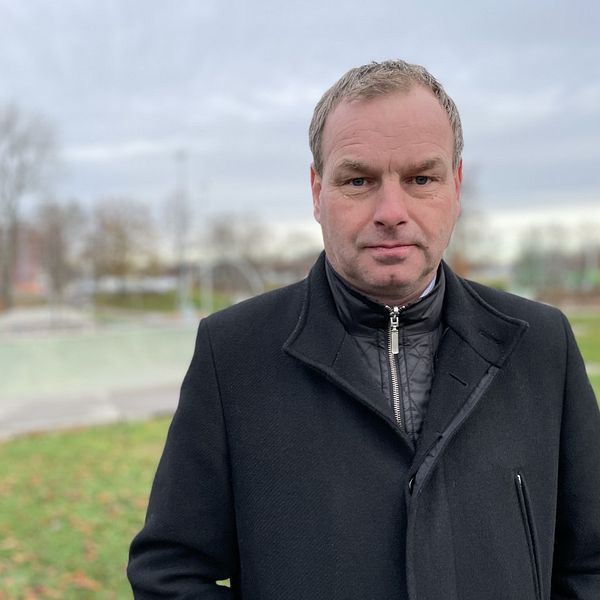 Person i mörk rock står utomhus, med träd och gräsmatta i bakgrunden.