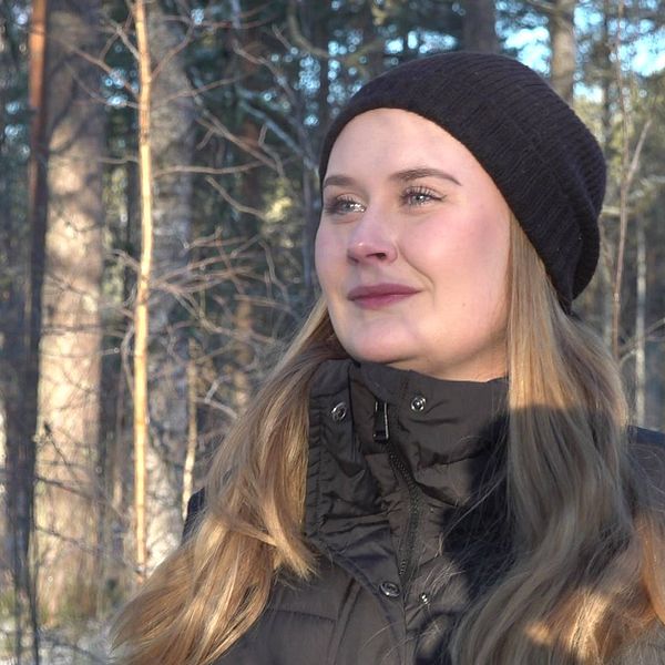 Bild på kvinna med svart mössa som står i en skog. Kvinnan heter Maja Johansson och ska i sitt examensarbete undersöka hur Storforsborna ser på skogen.