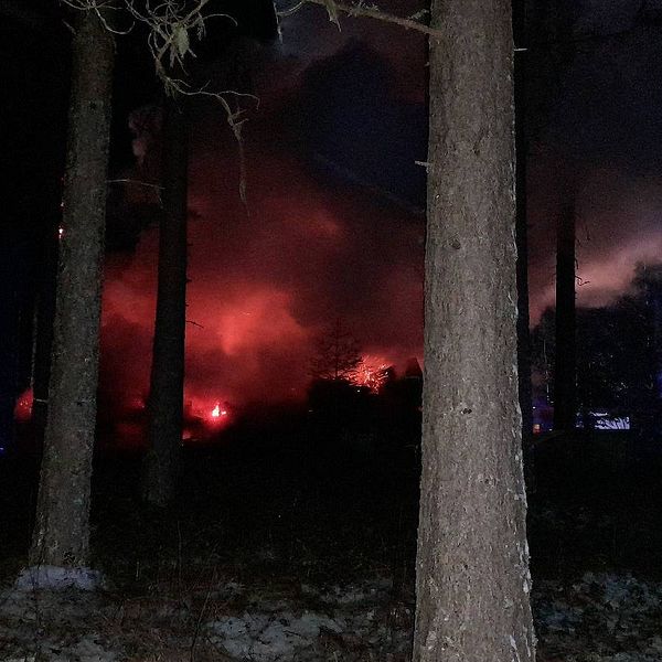 När räddningstjänsten kom till platsen var huset övertänt. På bilden ser man en brand på avstånd i mörker. I förgrunden syns träd och brandmän.