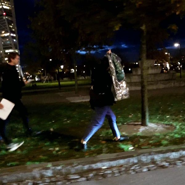 Två av SVT:s reportar följer efter Soheil Naderi i en park i Malmö på kvällen. Turning torso syns i bakgrunden.