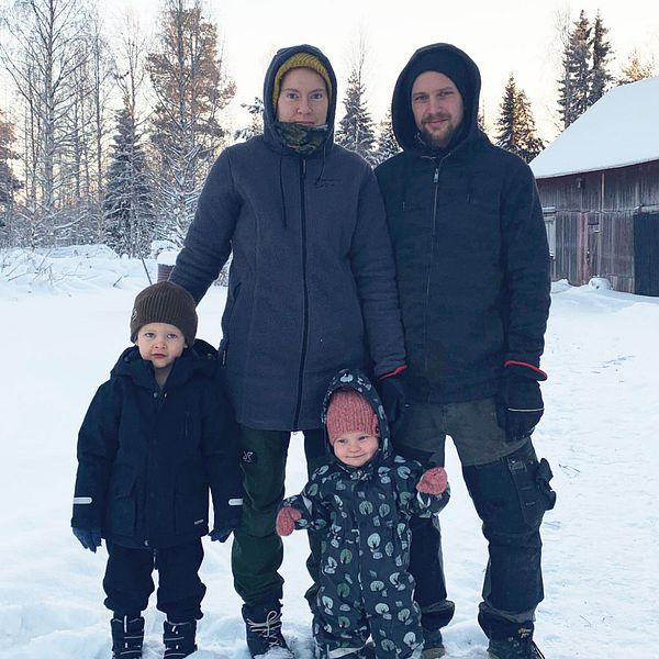 En mamma, pappa, son och dotter. I bakgrunden ser man en ladugård och ett gammalt timmerhus. Det ser kallt ut och det är mycket snö på marken.