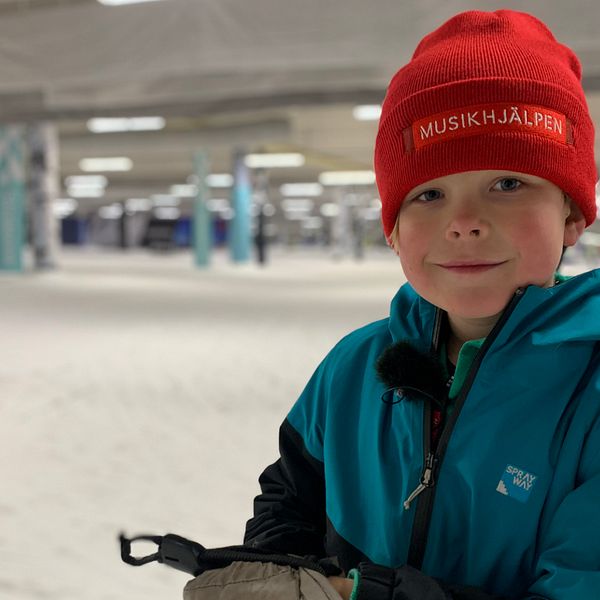 Bild på en pojke med röd mössa där det står Musikhjälpen. Pojken heter Arvid och står i en anläggning för längdskidor där han ska åka för att samla ihop pengar.