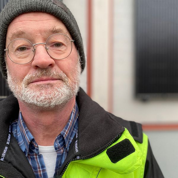 Porträttbild på Per Ekstorm, energi- och klimatrådgivare i Eskilstuna kommun. Han har på sig varseljacka, mössa och runda glasögon. I bakgrunden skymtar två solcellspaneler.