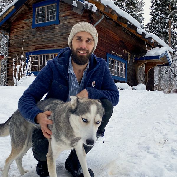 Youtubern Kalle Flodin sitter på huk i snön och håller sin huskey Tuss i famnen. I bakgrunden ser vi hans timmerstuga.