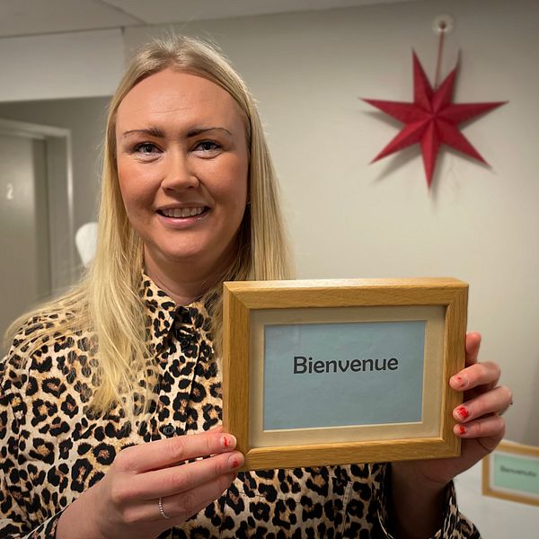 Bild på en blond kvinna med en skylt där det står Bienvenue. Kvinnan heter Elina Perdsjö och är verksamhetsledare för natthärbärget Vinternatt. Hon bjuder på en rundtur i de nya lokalerna.