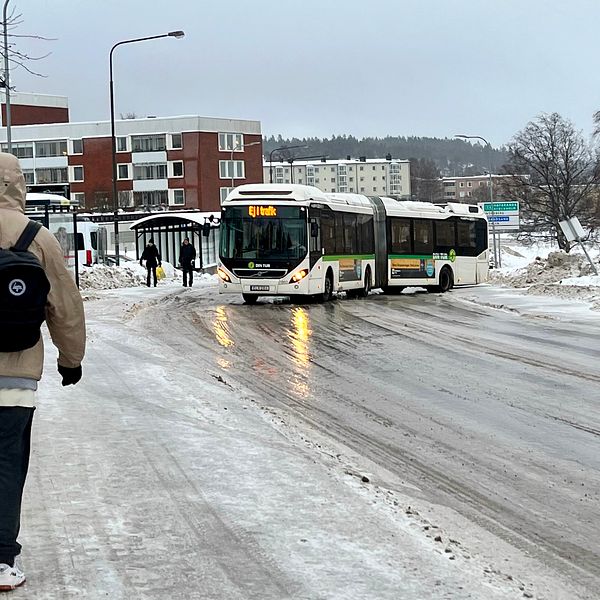 En man går med en svart jacka på en hal trottoar med två blåa soppåsar i handen. Till höger om honom slirar en buss som har glidit rakt över vägen på grund av halkan i Sundsvall.