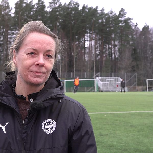 Kubbchef för IFK Värnamo Lisa Lidén lyssnar på intervjufrågan. I bakgrunden tränar klubbens gymnasielag på konstgräsplanen.