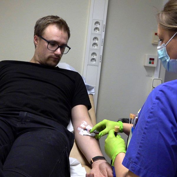 Bild på en man med svart t-shirt och svarta byxor som ligger på en sjukhussäng. Bredvid honom sitter en läkare med blå tröja och som håller i mannens omplåtstrade arm. Mannen heter Oliwer Andersson och är patient inom ett forskningsprojekt om diabetes.