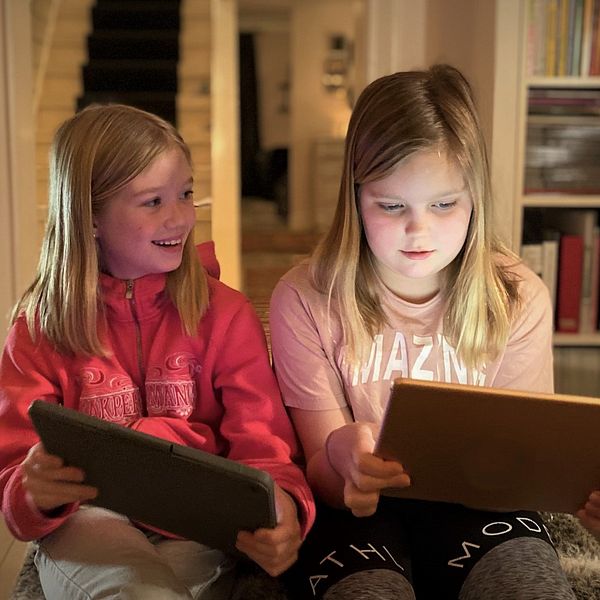 Två flickor sitter med varsin tablett på vardagsrummet och spelar dataspel på plattformen Roblox