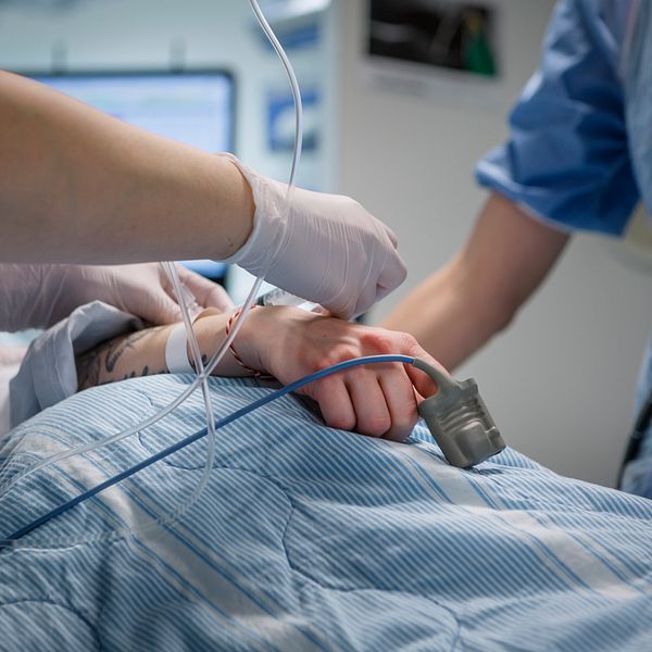 Bilden visar en sjukhussäng där man ser en hand på ett täcke, med en syremätare på fingret, samt två personer ur vårdpersonal som hjälper patienten. Danderyds sjukhus.