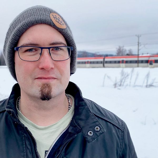 En man med mössa och glasögon tittar in i kameran. I bakgrunden ett tåg i ett snölandskap.