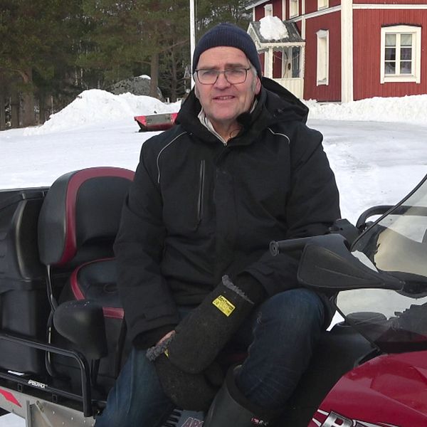 Johnny Stålarm bor på Hindersön i Luleå skärgård. Han sitter iklädd svart mössa och svarta vinterkläder på en djupröd skoter. I bakgrunden syns snö, barrskog och ett rött hus med vita knutar.