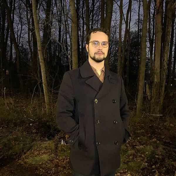 En helbild på Martin Eriksson, glasögon, skägg, mörkt hår, som står framför en mindre skogsdunge i eftermiddagsmörkret
