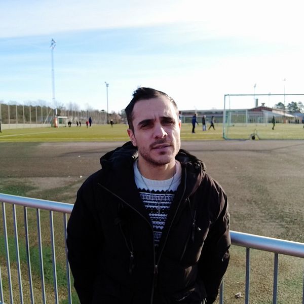 Fotbollsdomaren Sami Dalali står framför en fotbollsplan där spelare håller på med en träning.