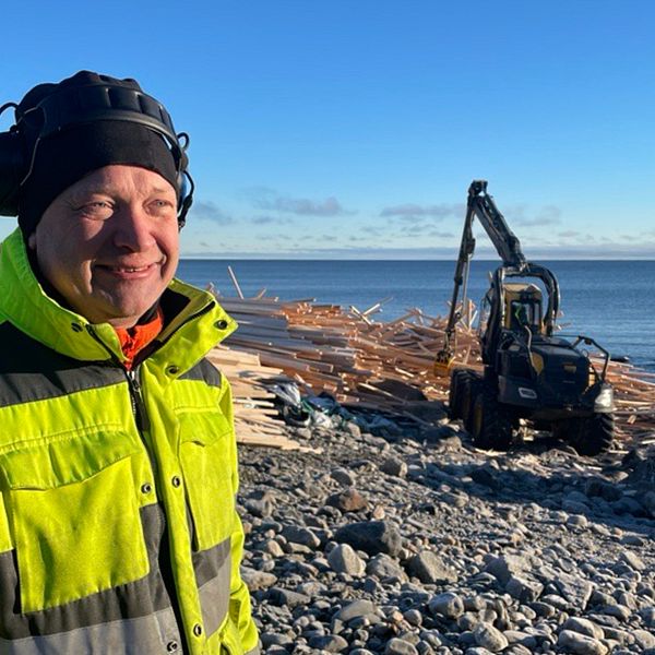 Mikael Danielsson som arbetar som bärgare står i arbetskläder ute vid kusten i Sjöviken utanför Härnösand. I bakgrunden syns en av maskinerna som plockar virket från vattnet och stranden. Virket ligger så långt ögat kan nå.