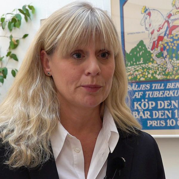 En kvinna (Majblommans generalsekreterare Åse Henell) Står i en korridor, bakom henne ginns en växt och en äldre affisch för majblomman, priset var 10 öre.