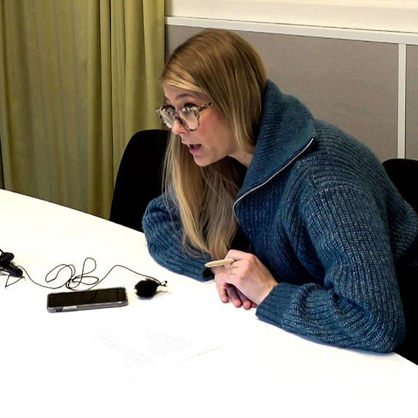 SVT:s reporter Fanny Asplund sitter vid ett vid stort bord med en telefon framför sig. Hon har öppen mun och pratar med någon. Hon tittar ut till vänster i bilden.