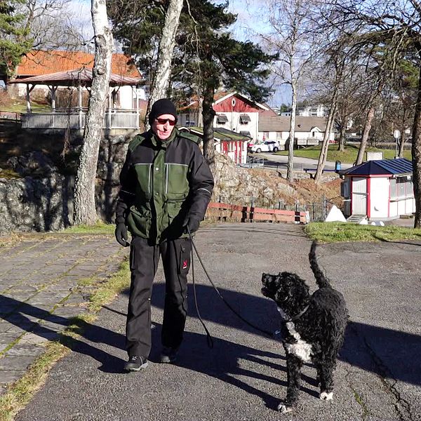 En äldre man med solglasögon och mössa går med en svart hund i koppel.
