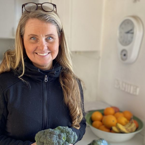 Det är en porträttbild av Malin Lindgren Sundström, som står i sitt kök och håller i en broccoli. Hon tittar och ler mot kameran.