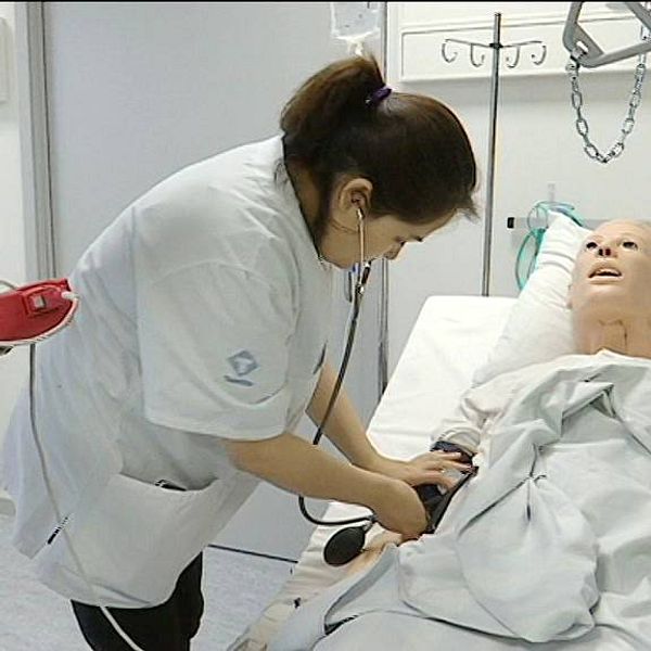 Kvinna med mörkt hår står böjd över en docka som ligger i en sjukhussäng. Kvinnan går sjuksköterskeutbilding på Mittuniversitetet i Östersund.