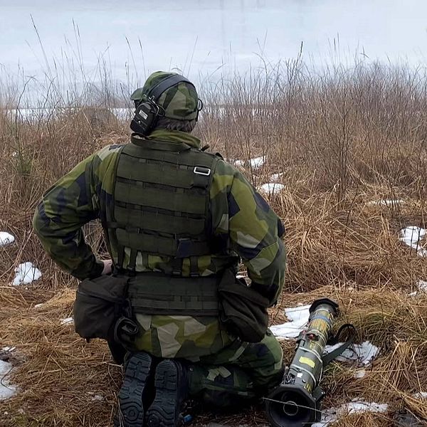 En soldat sitter på huk vid ett tomt fält med högt gräs och lite snö, med ryggen vänd mot kameran. På marken bredvid honom ligger en granatkastare.