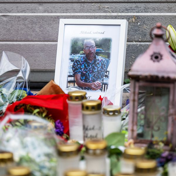 Ett fotografi av den mördade mannen Ulf Sandberg vid mordplatsen, med texten ”Älskad, saknad” och en stor mängd blommor och ljus.