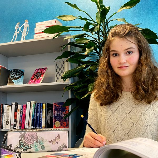13-åringen Emilia Kenaga sitter vid ett arbetsbord med en stor grön växt bakom sig och har skolböcker framför sig och en penna i handen.