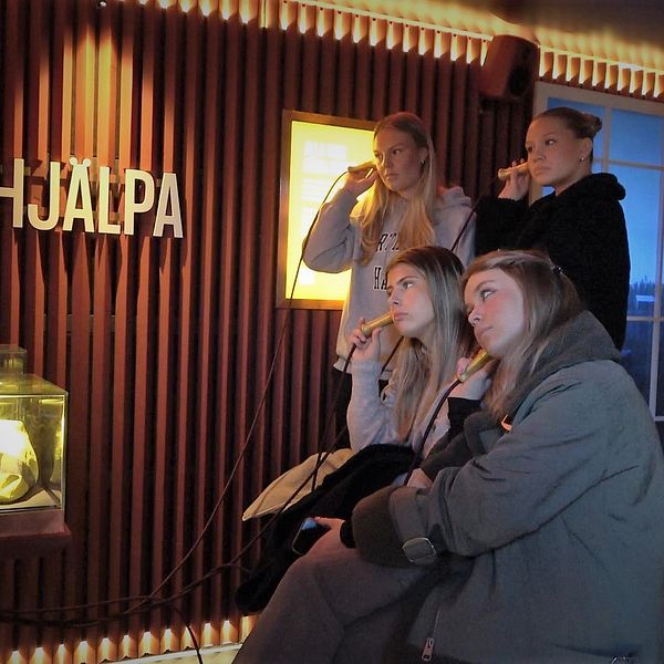 Fyra tjejer håller varsin hörlur mot örat inne i en rödmålad monter och blickar uppmärksamt mot vänster. Ordet ”Hjälpa” står upphängd på väggen.