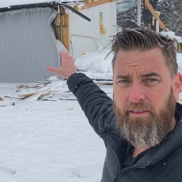 Vd:n Antony Viklund, står inför det raserade taket och pekar mot den del av byggnaden som består. Viklund har en svart jacka och skägg och står i ett snöigt landskap i Robertsfors kommun.