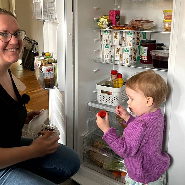 Klara Twete sitter på huk framför kylen och ger sin dotter en jordgubbe.