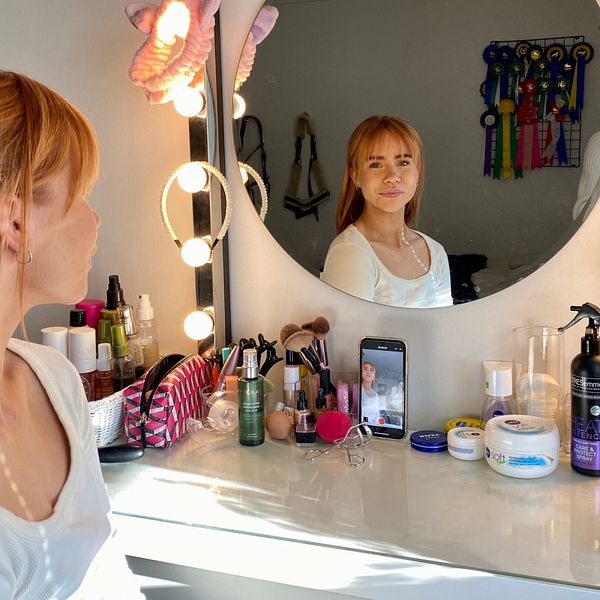 Influencern Ullabritta sitter vid sitt sminkbord och tittar in i kameran via spegeln. På sminkbordet ligger hennes mobil som spelar in en video.