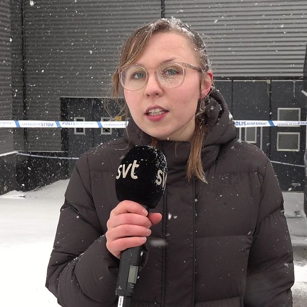 Bild på SVT:s reporter Serafia Olausson som håller i en SVT-mikrofon. Bakom henne syns polisavspärrningarna.